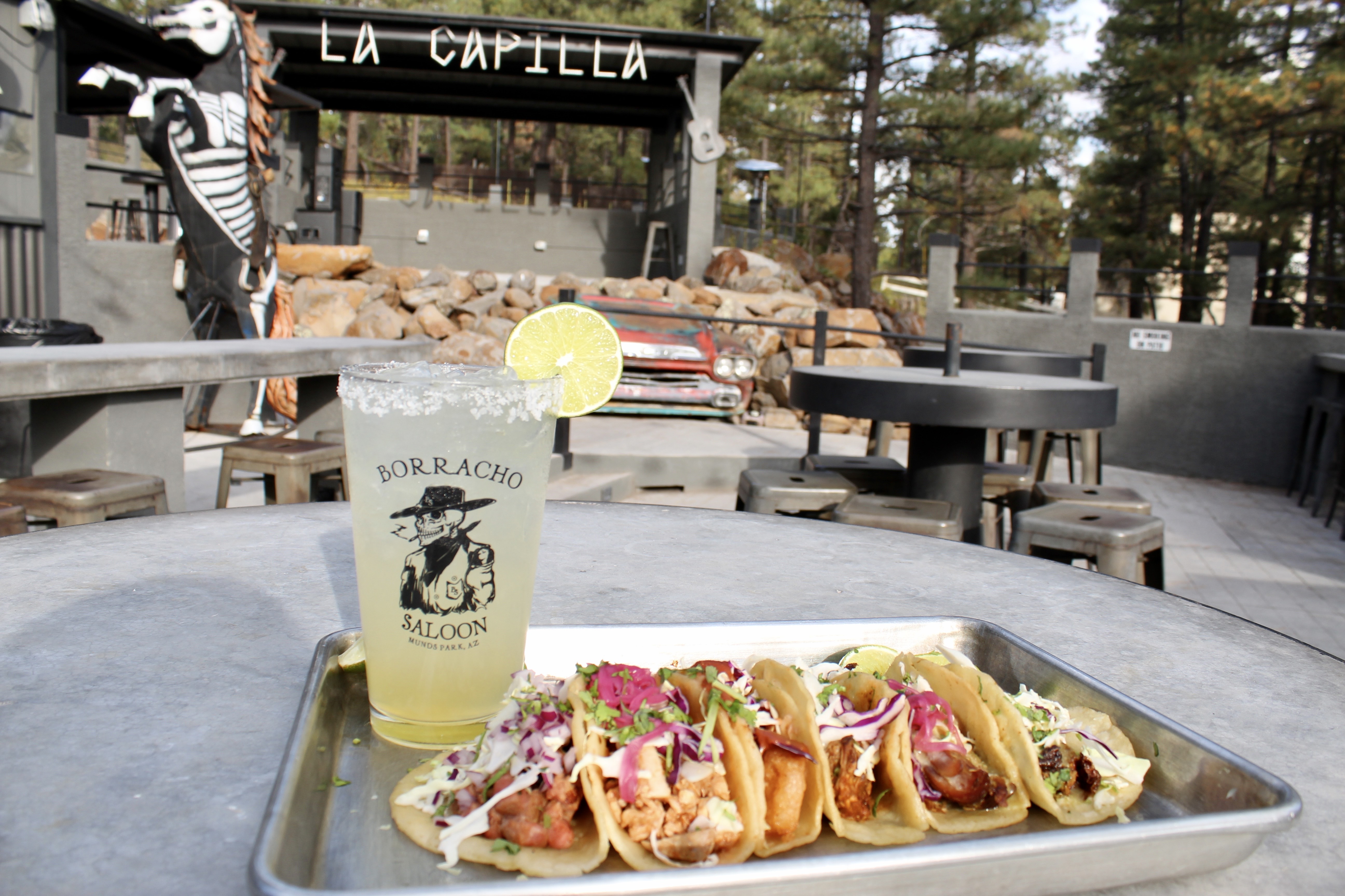 Borracho Saloon tacos of various meats plated on a silver tray with a margarita in a Borracho Saloon glass and a view of the Borracho Saloon La Capilla stage