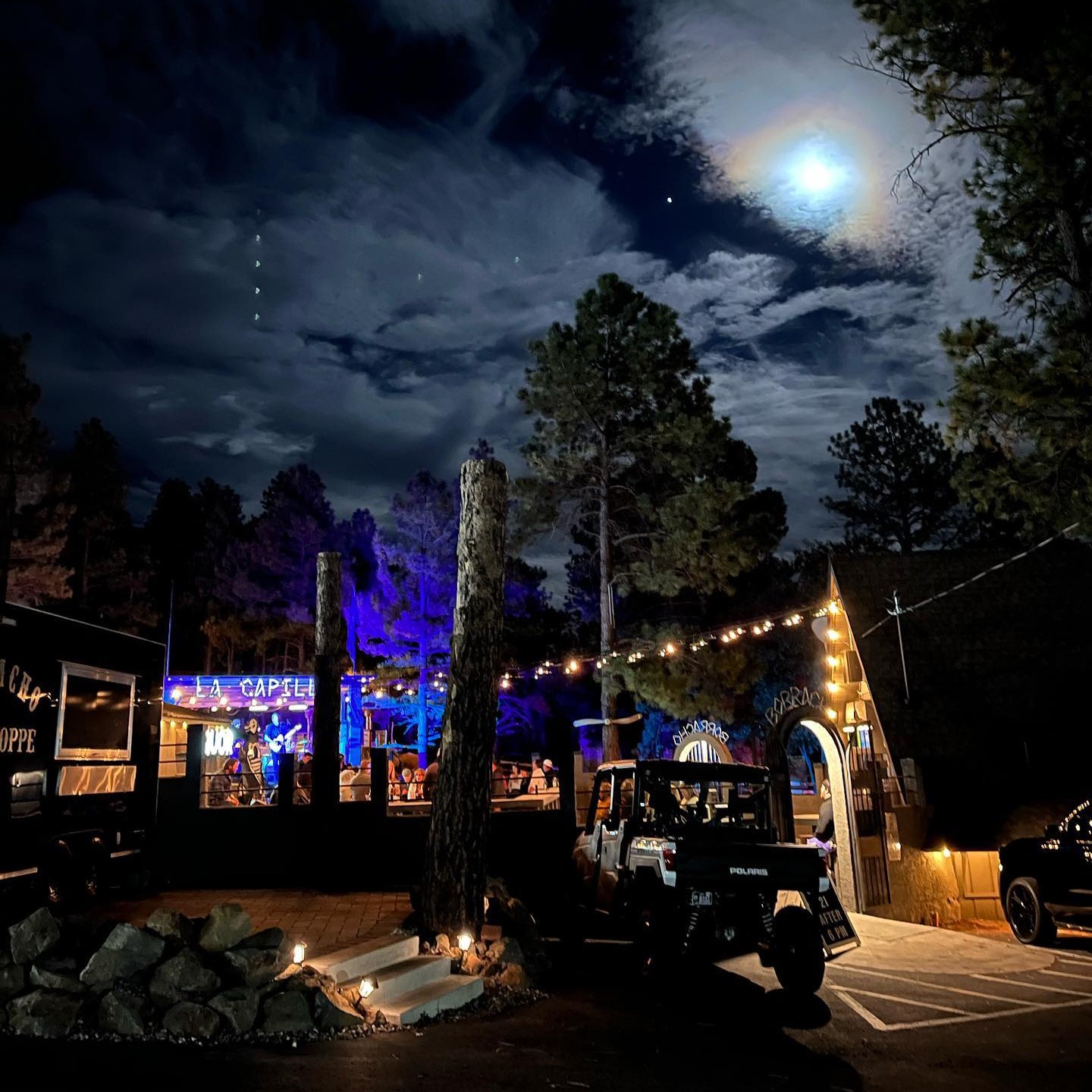 View from the Borracho Saloon parking lot with the Taco Shoppe on the left, the La Capilla stage and large trees in the middle background, Borracho Saloon itself on the right, and a bright moon illuminating the sky