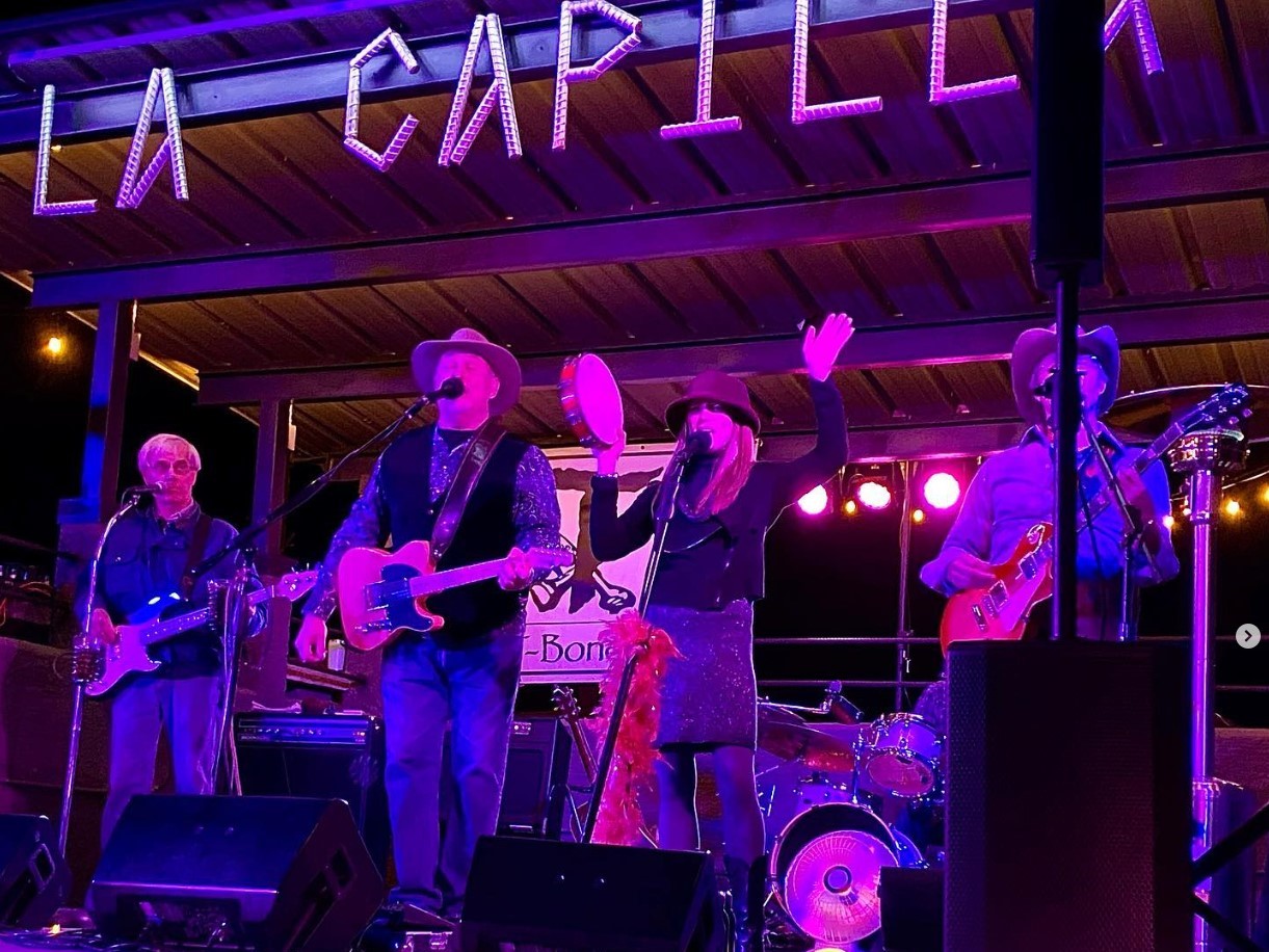 3 men singing playing guitar and a woman singing playing the tamborine on the Borracho Saloon La Capilla stage